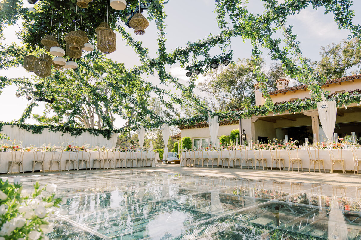 Outdoor wedding ceremony space with a large glass dance floor. The space is decorated with lots of ivy hanging from wire frame above.