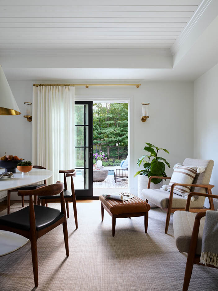 Discover the casual dining room in a historic Hingham home, featuring a new sliding door with views to the patio and pool, a custom bench and table, white linen drapes, and mid-century furnishings.