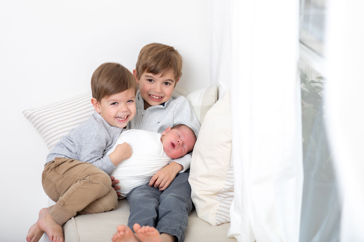two sibling brothers hold their newborn baby sibling who is swaddled and sleeping, the brothers smile.