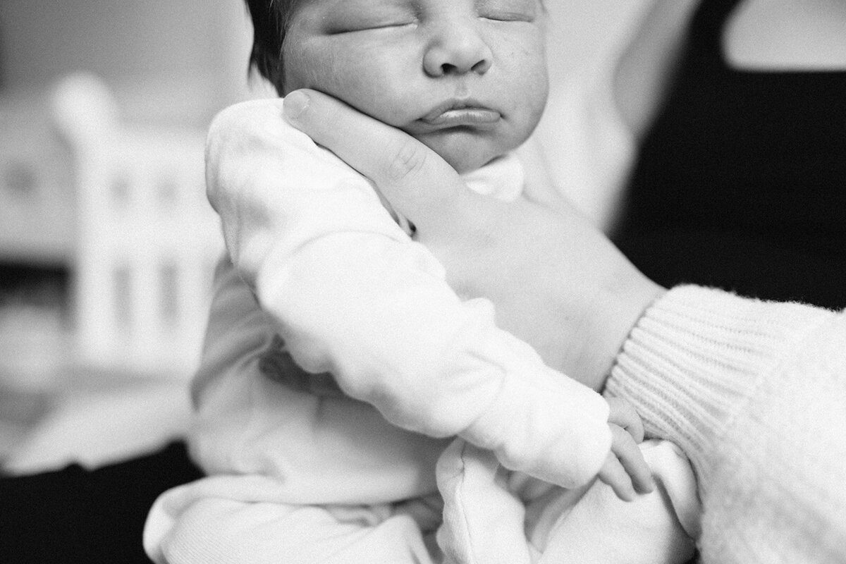 Close-up of a newborn baby in white clothing, held gently by an adult's hand, eyes closed. Captured in stunning detail by a Toronto newborn photographer. Black and white image.