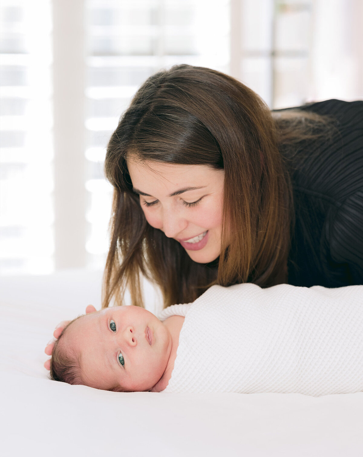 Mother smiling over her newborn baby swaddled in white during an in-home lifestyle session by Isela Lozano, San Francisco newborn photographer serving families in Noe Valley, Palo Alto, Menlo Park, Marin, and the Bay Area.