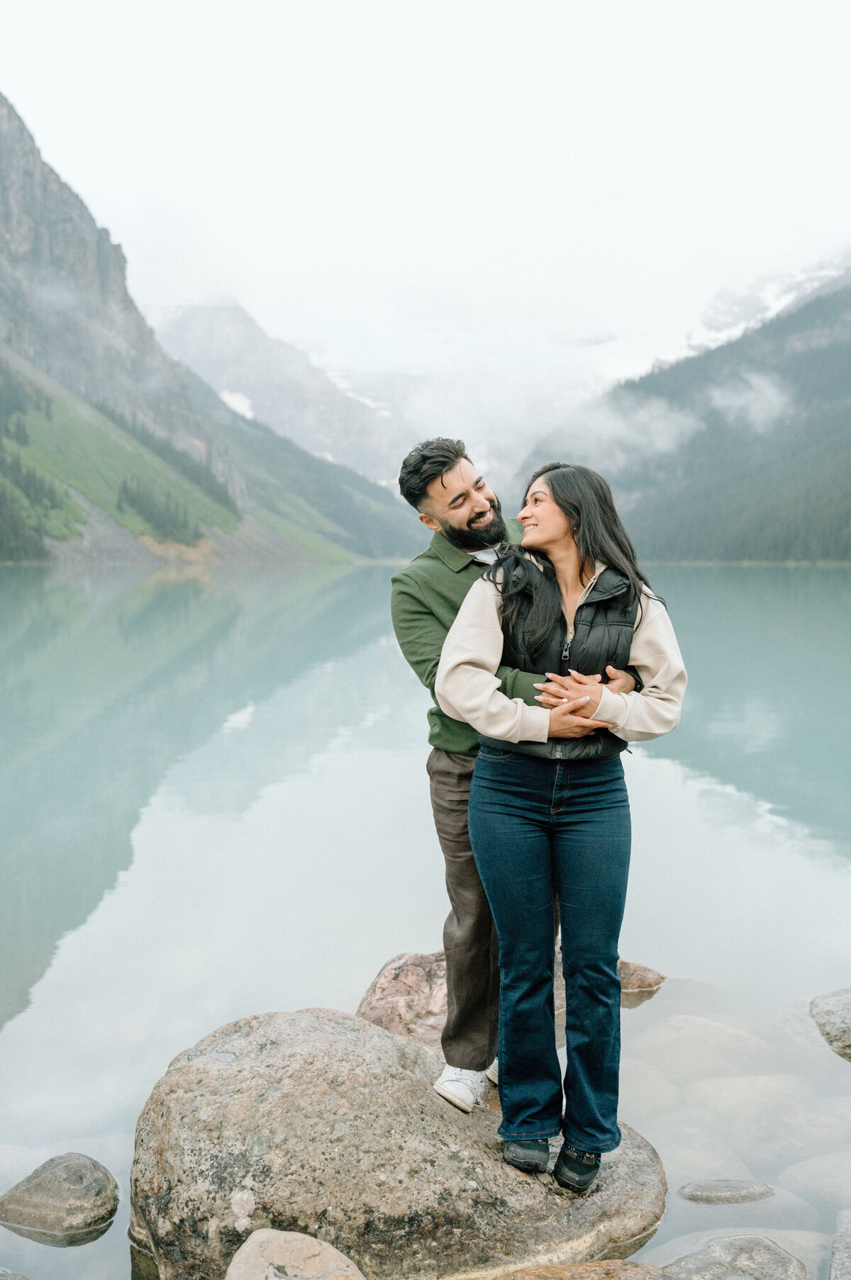 Sweet hug at Lake Louise during engagement session with misty mountains and calm water.