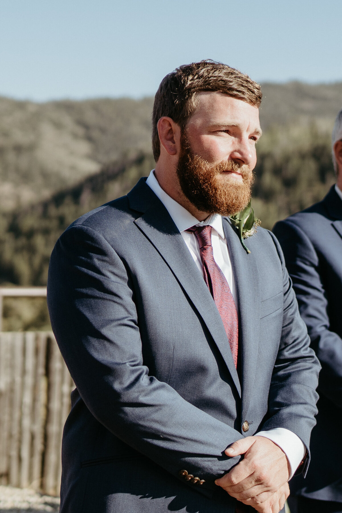 Groom in a navy suit and burgundy tie waits for his bride on his wedding day.