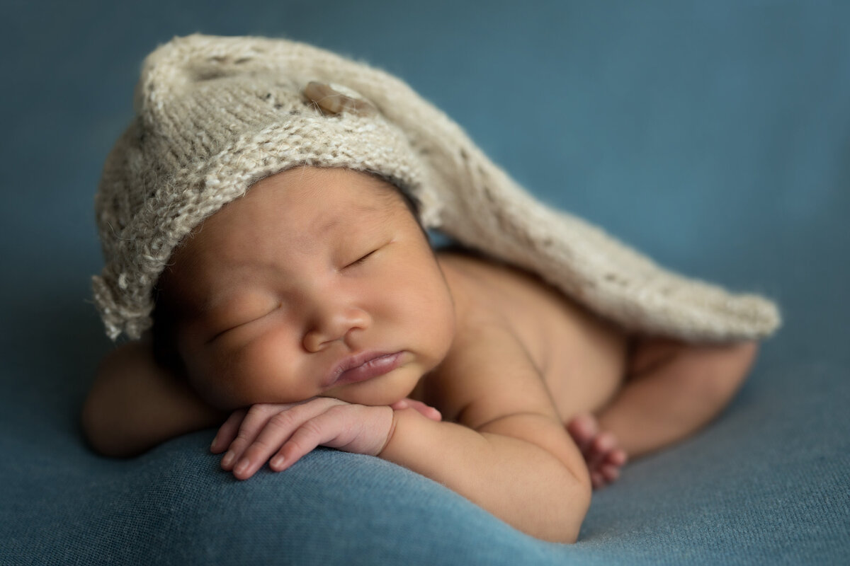 Asian baby with long, knit cream colored hat.  He is laying on his arms and his feet are curled under him.  He is on a blue backdrop.
