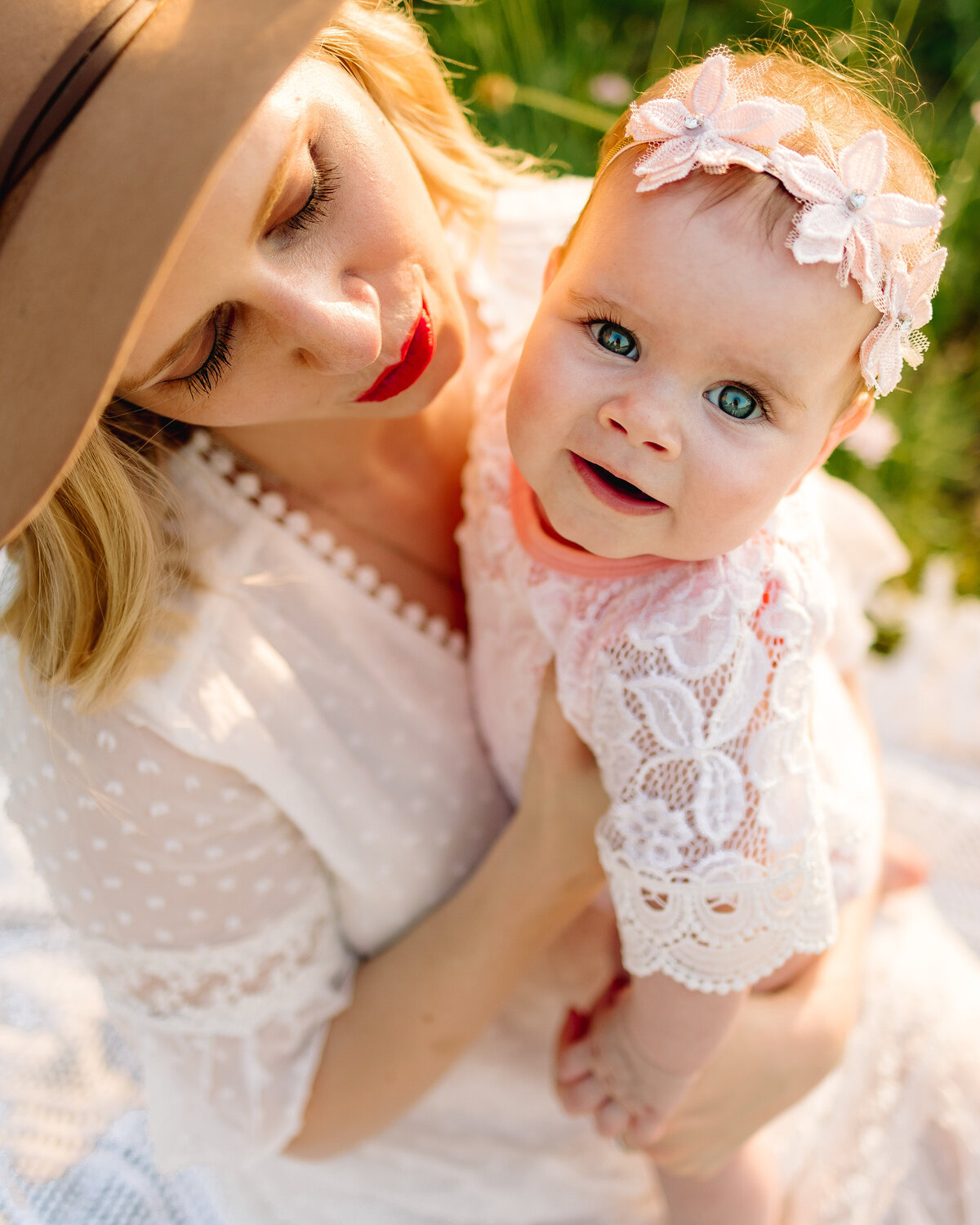 Picture of a woman with a baby, she is watching her she has a brown hat and red lips is holding the baby looking at the camera. The baby has blue eyes and on her head has a headband with light pink flowers
