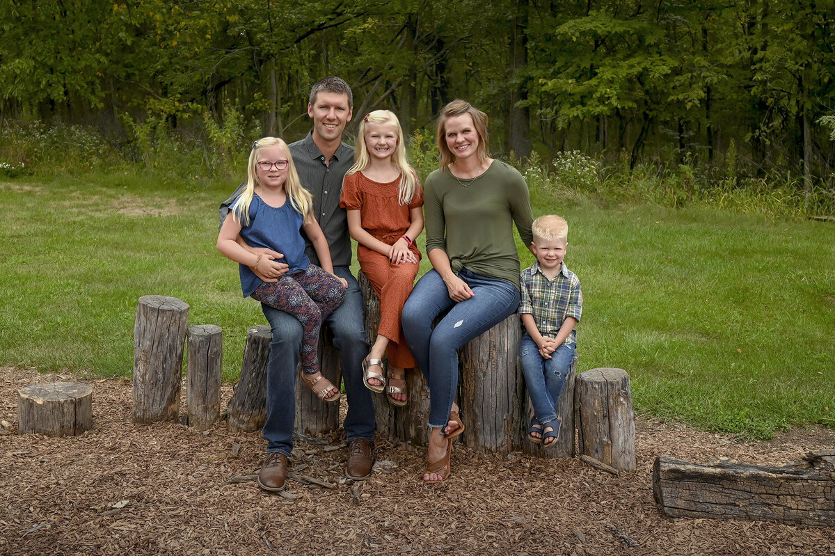 Family of four sitting on wooden logs outdoors, smiling with trees in the background.