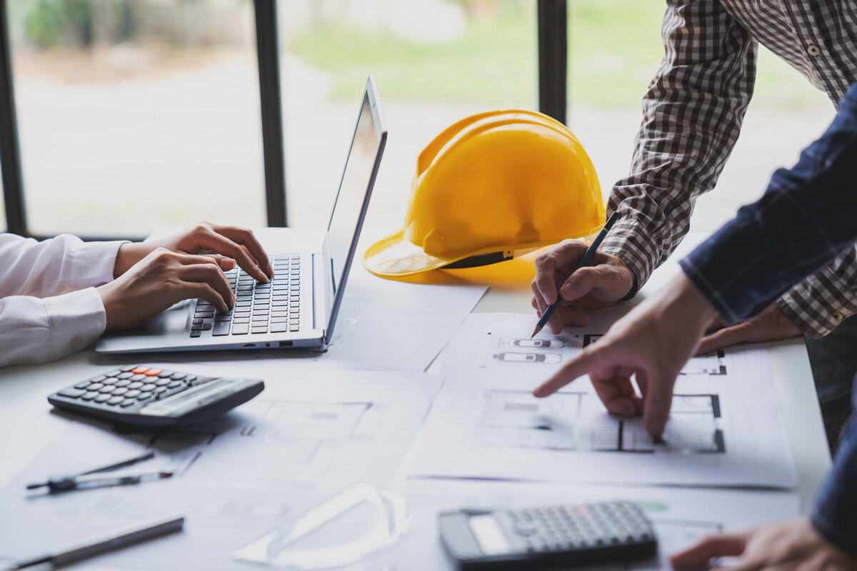Custom home builders working on blueprints at a desk with a laptop, calculator, and yellow safety helmets, emphasizing collaboration in design and construction.