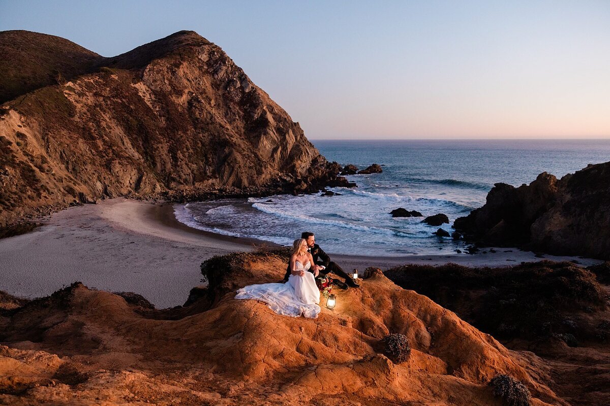 A bride and groom sit on the cliff edge during sunset overlooking the ocean in Big Sur.