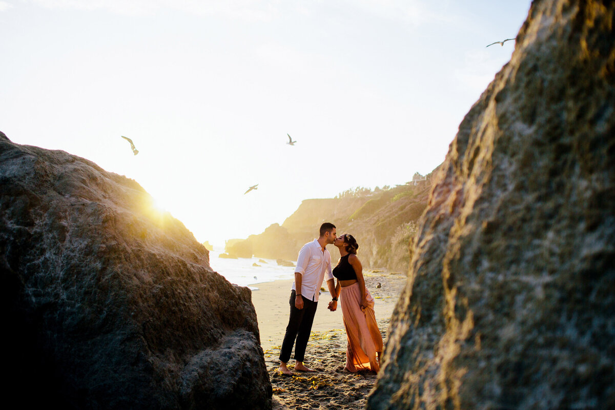 Man and woman kiss on a rocky beach during the sunset