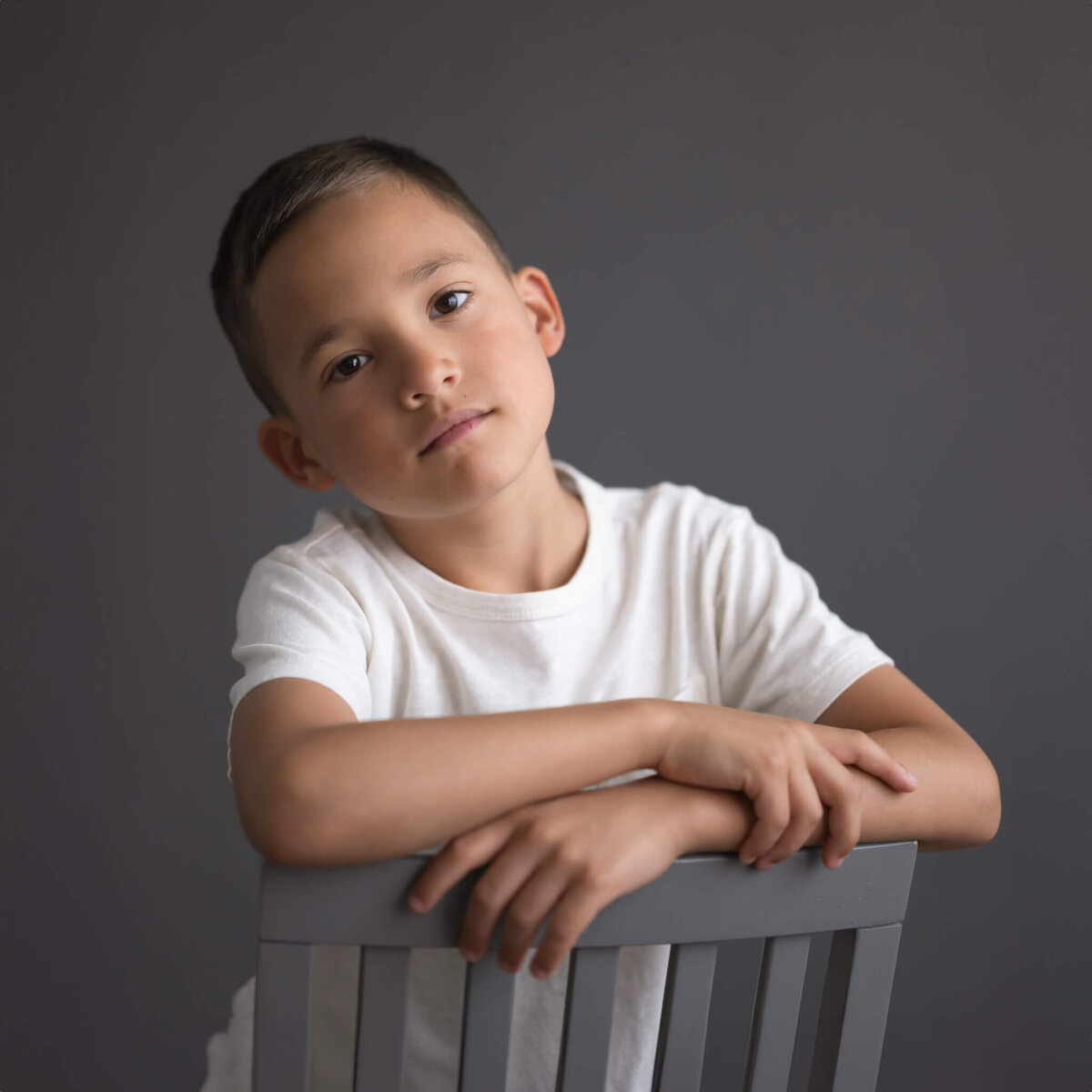 gorgeous studio portrait of 8 year old boy in white tee shirt sitting backwards on chair with grey background and a serious face