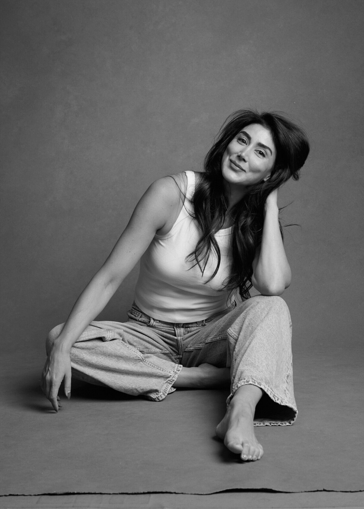 Black and white portrait of a woman sitting barefoot on the floor, wearing a white tank top and relaxed-fit jeans. She leans on one hand with a soft smile, exuding confidence and ease. A natural, unfiltered moment from 'THIS IS ME – A Photography Experience by Katie Campbell' in Atlanta, GA, celebrating authenticity and self-love.