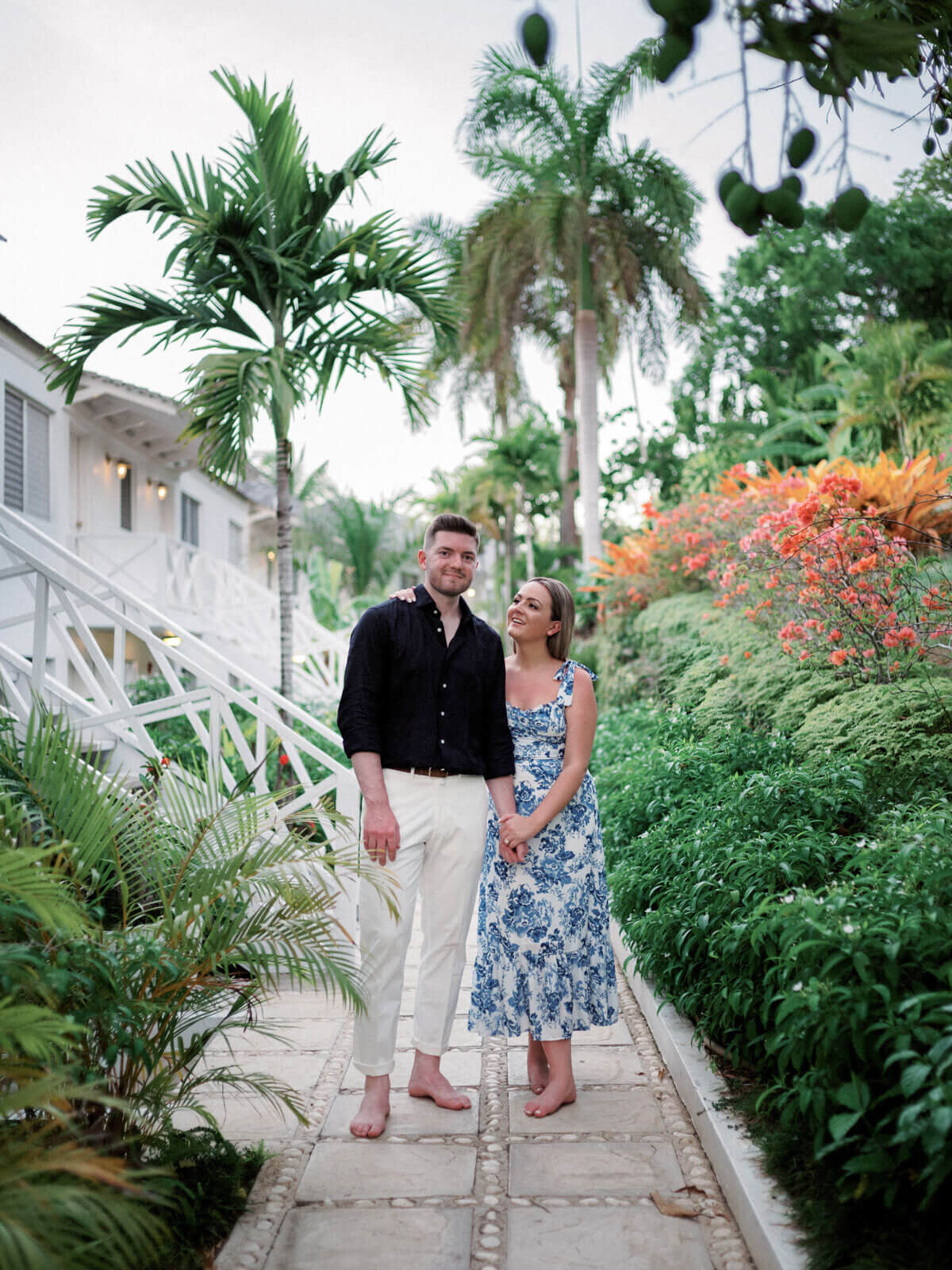 The engaged couple is standing on a pathway, in the garden of Round Hill Hotel and Villas, Jamaica.
