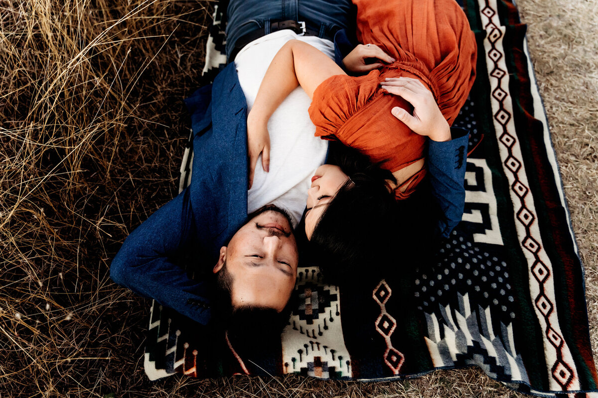 Couples Photography, Man laying on his back wearing a blue blazer as woman in a rust dress lays on top of him snuggling. They are both laying on a tribal patterned blanket.
