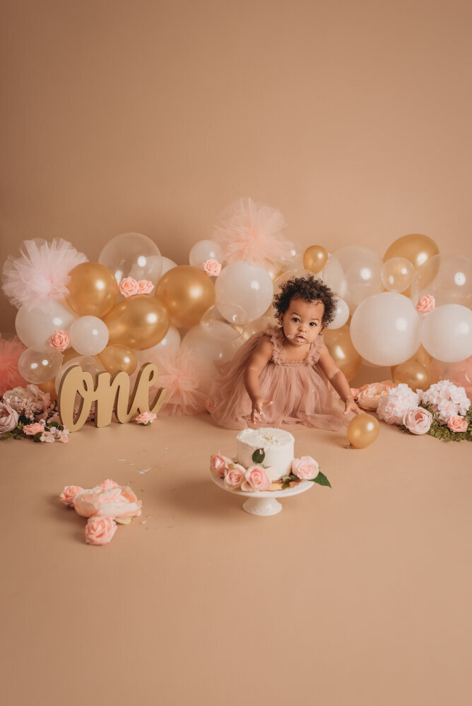 Cake smash session with Atlanta, Georgia cake smash photographer of one year old baby girl in pink tulle dress on tan background with neutral colored balloon garland eating her white iced birthday cake with pink roses on it