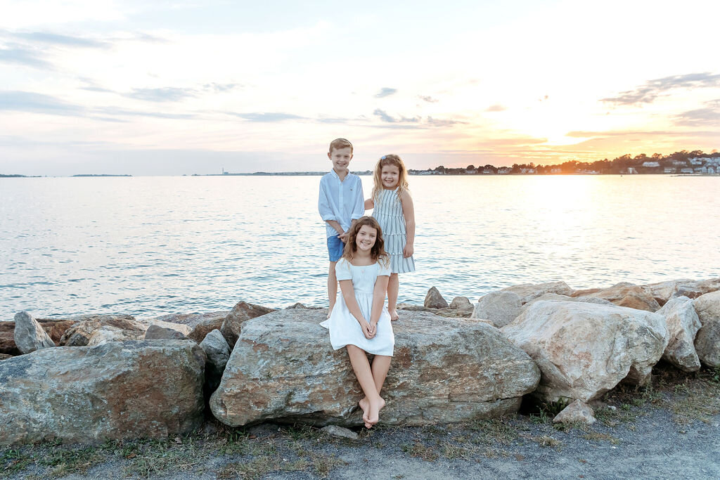 Three siblings posed on coastal rocks during sunset session photographed by Connecticut family photographer at beach location