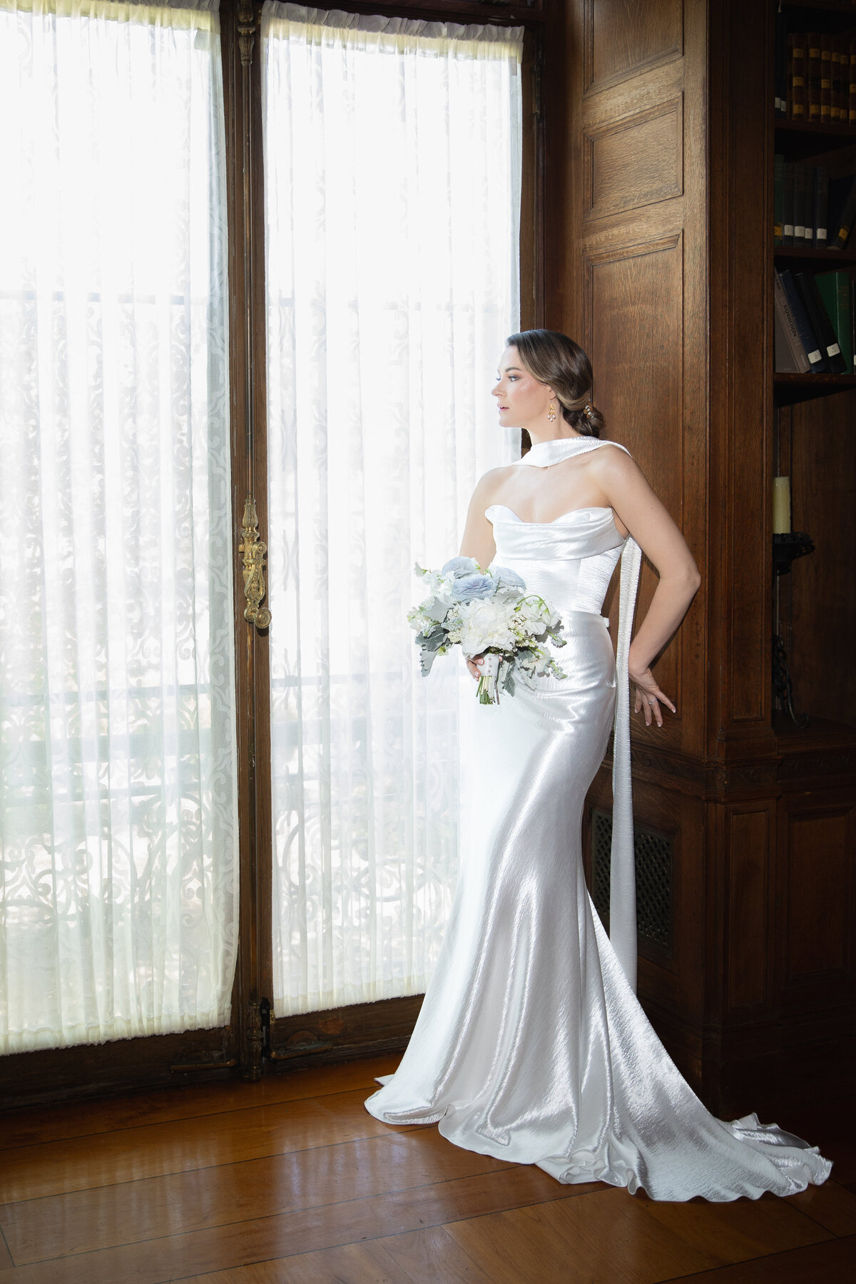 Rhode Island Wedding Photographers | Kelsey Sheehan Photography Timeless Rhode Island Weddings | A woman stands by a window wearing a shiny, elegant white gown, holding a bouquet of white and green flowers. The mood is serene and sophisticated.