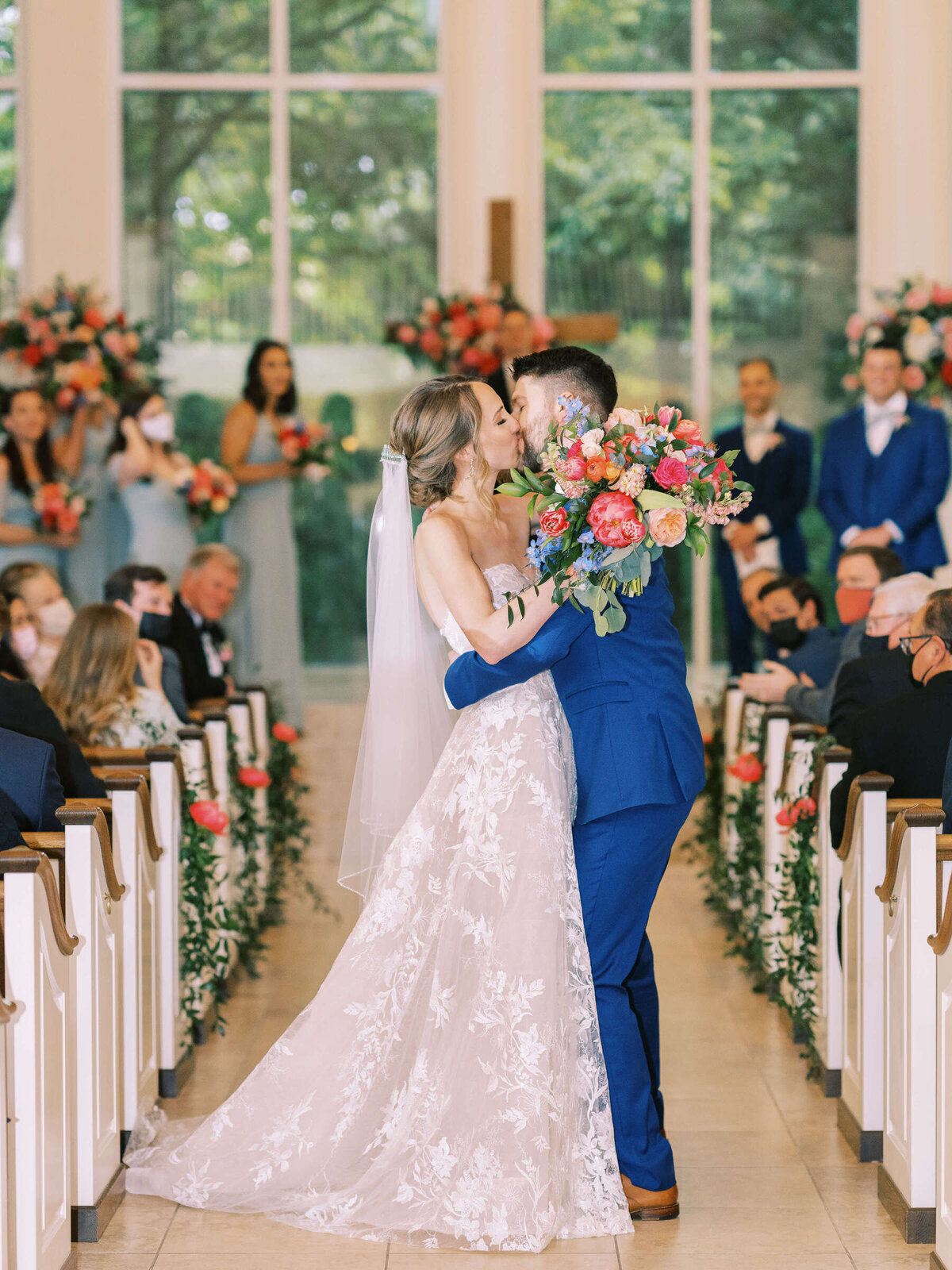 Bride and groom kiss in wedding chapel aisle with colorful bridal bouquet