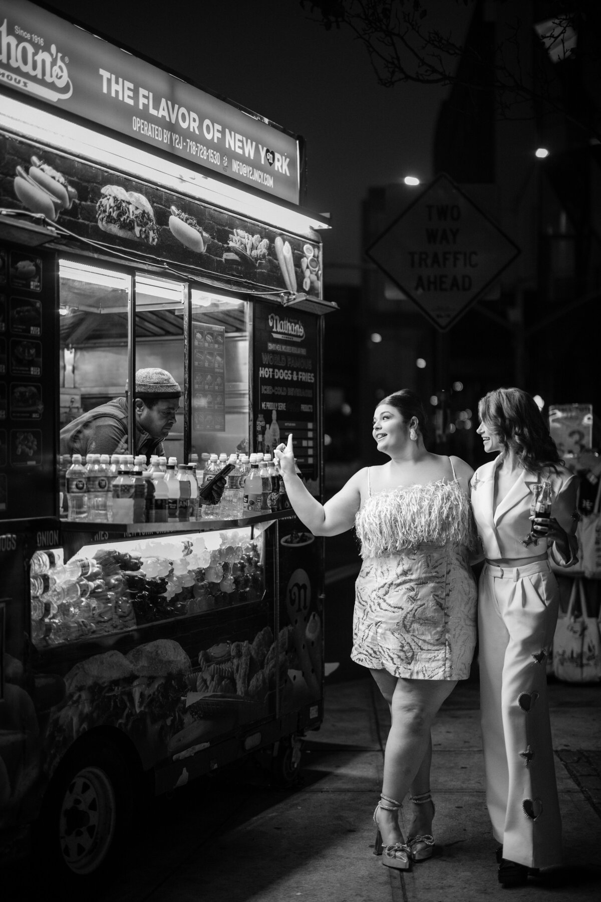 Two women at their NYC city street at night engagement photos session stand at a New York hot dog cart at night. One is ordering while the other holds a drink. The vendor is preparing food inside the cart.