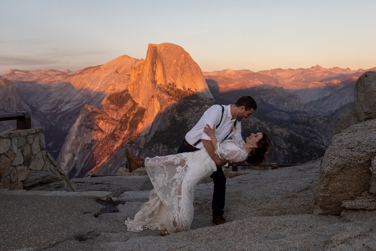 A groom dips his bride on their Yosemite elopement day as the sunsets and casts a pink glow over Half Dome behind them.