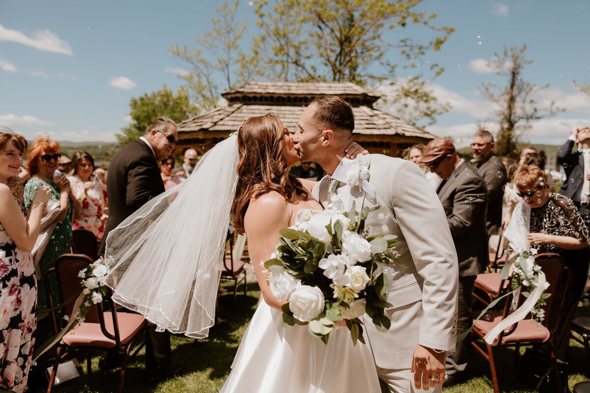 Bride and groom kiss during their joyful wedding ceremony exit at an outdoor Westchester wedding, surrounded by guests blowing bubbles under a sunny blue sky.