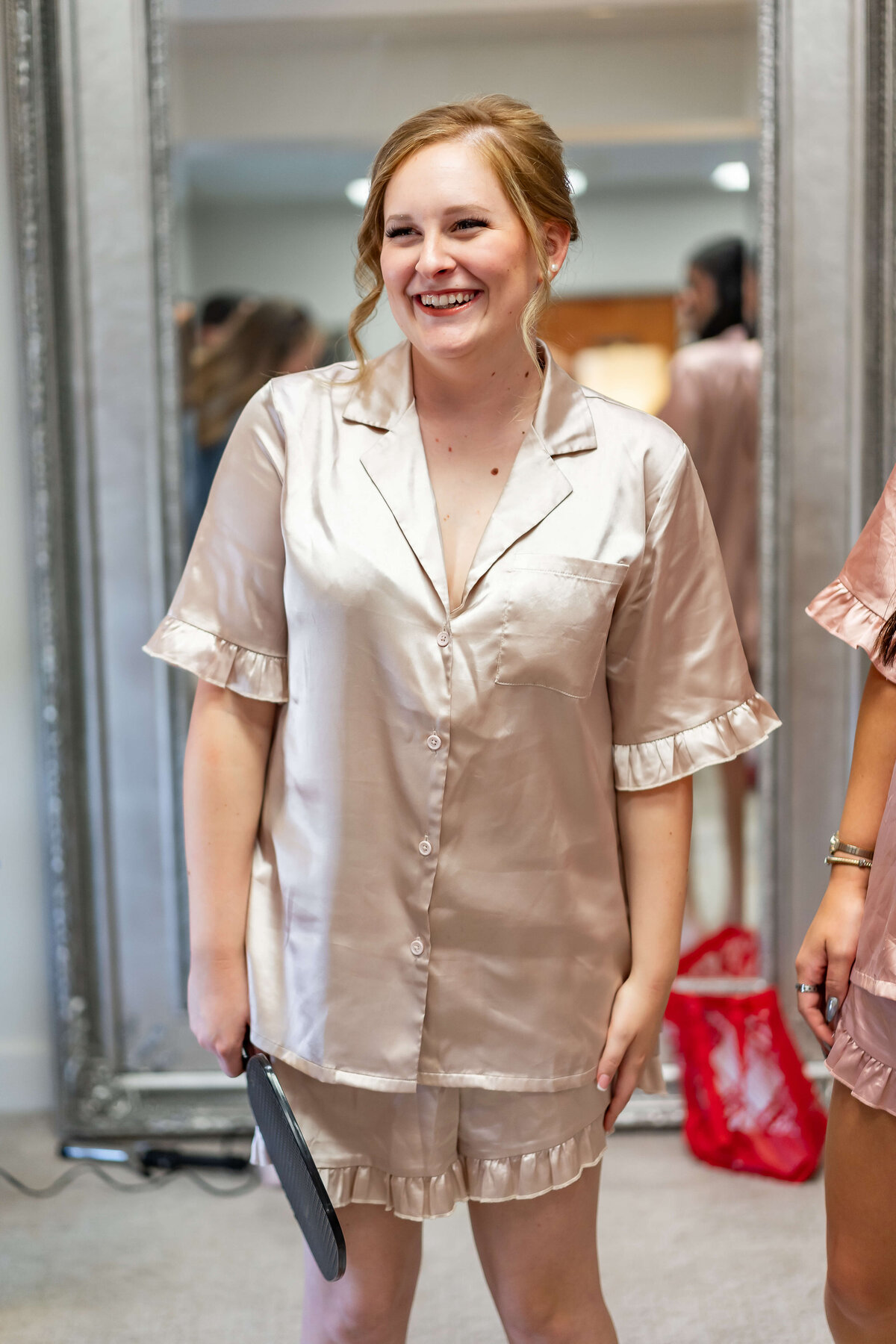 A woman in satin pajamas smiles while standing in front of a large mirror, with two other women in matching outfits in the background. The scene appears casual and joyful, likely during a pre-wedding event.