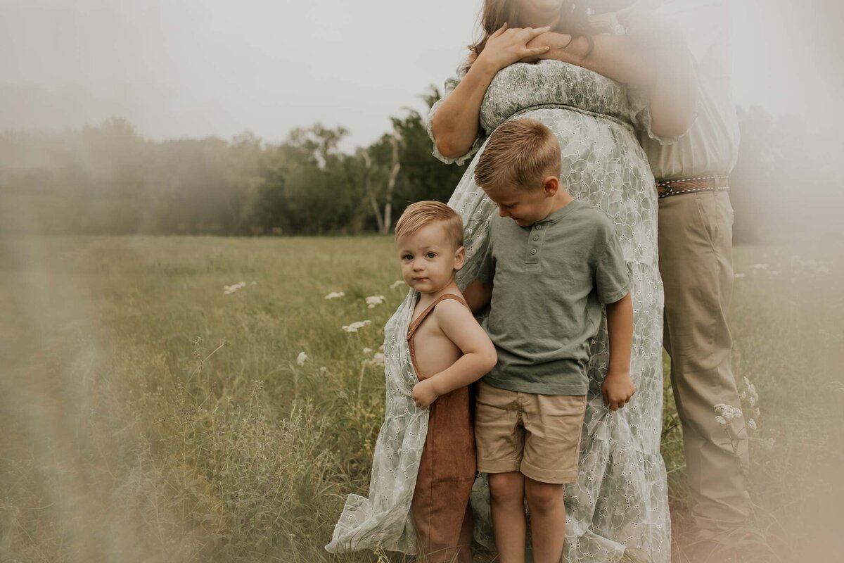In a field, young boy in overalls looks into the camera as his brother looks down on him, and their mother and father are embracing behind them.