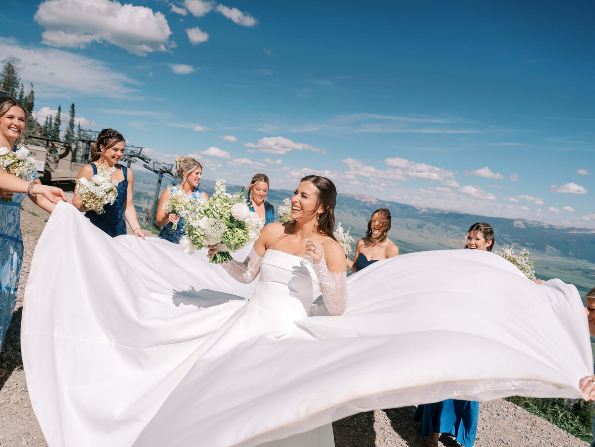 Bride laughing and twirling in her wedding dress surrounded by bridesmaids holding bouquets, with mountain views and blue sky in the background