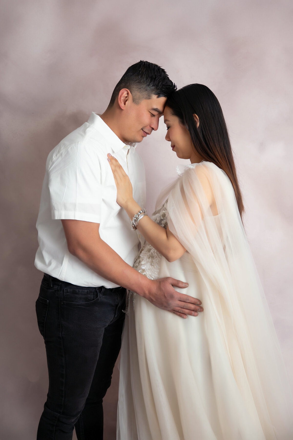 Expecting parents hug closely with foreheads touching, his hand on her belly and her hand on his chest.