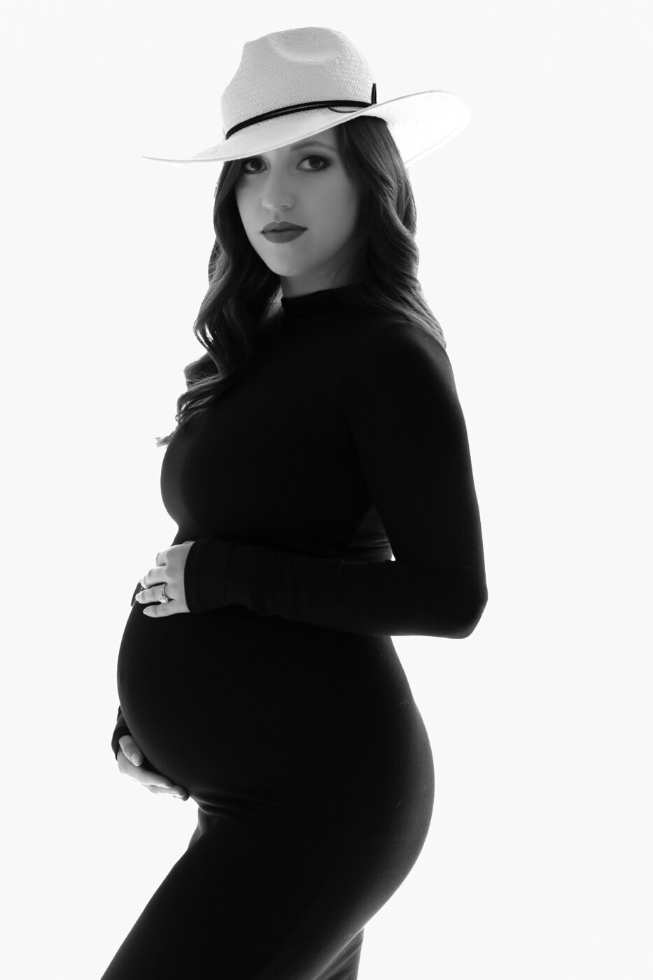 A woman posing on her side profile wearing a hat and a black dress showing her pregnant belly