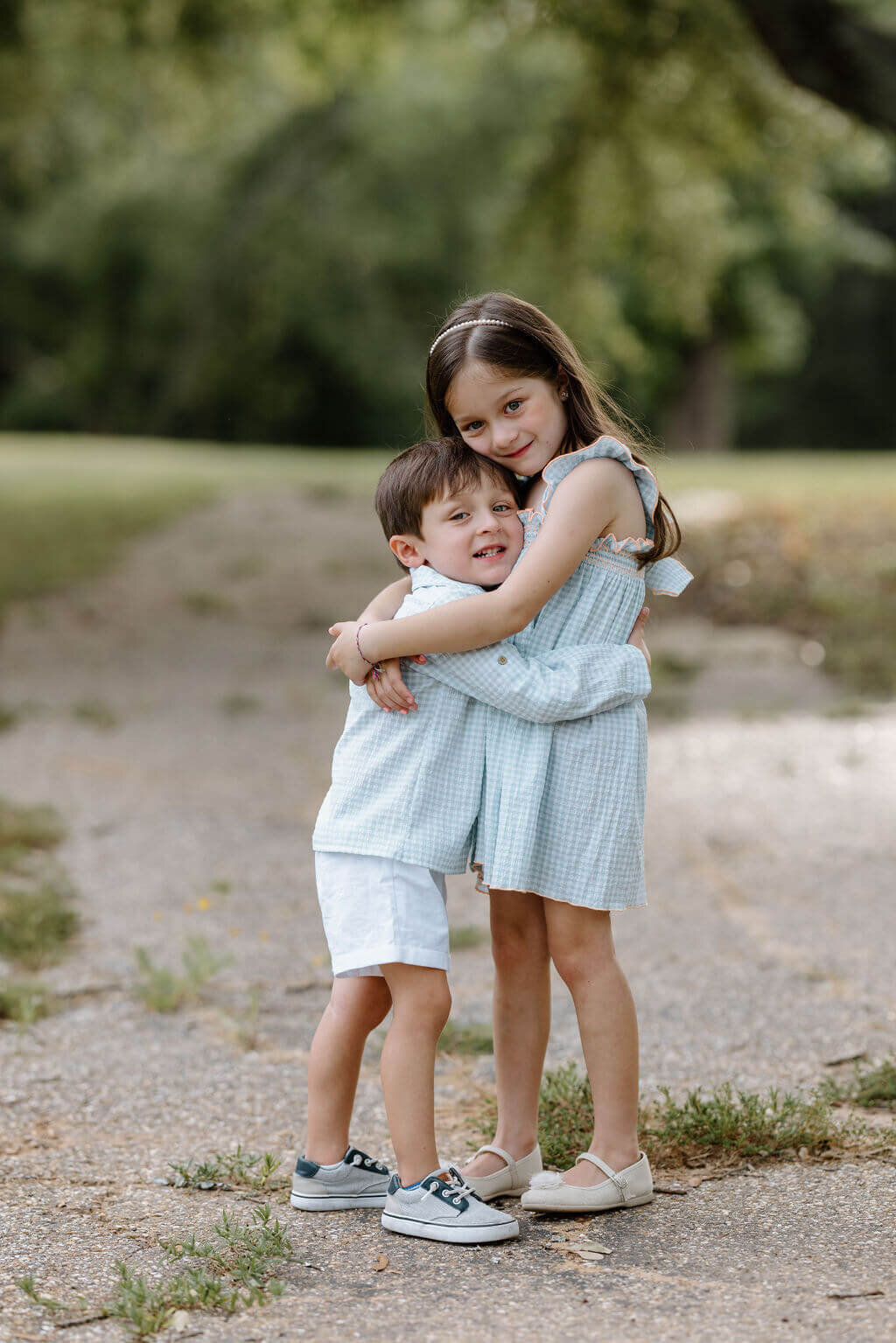 candid picture of two siblings in neutral summer clothing hugging under large oak tree