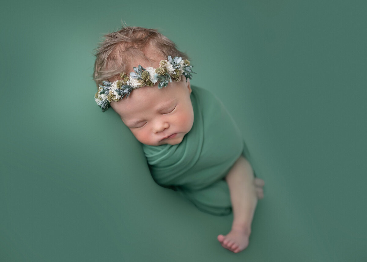 A newborn baby sleeps in a floral headband and green swaddle in the studio of a Pella Newborn Photographer