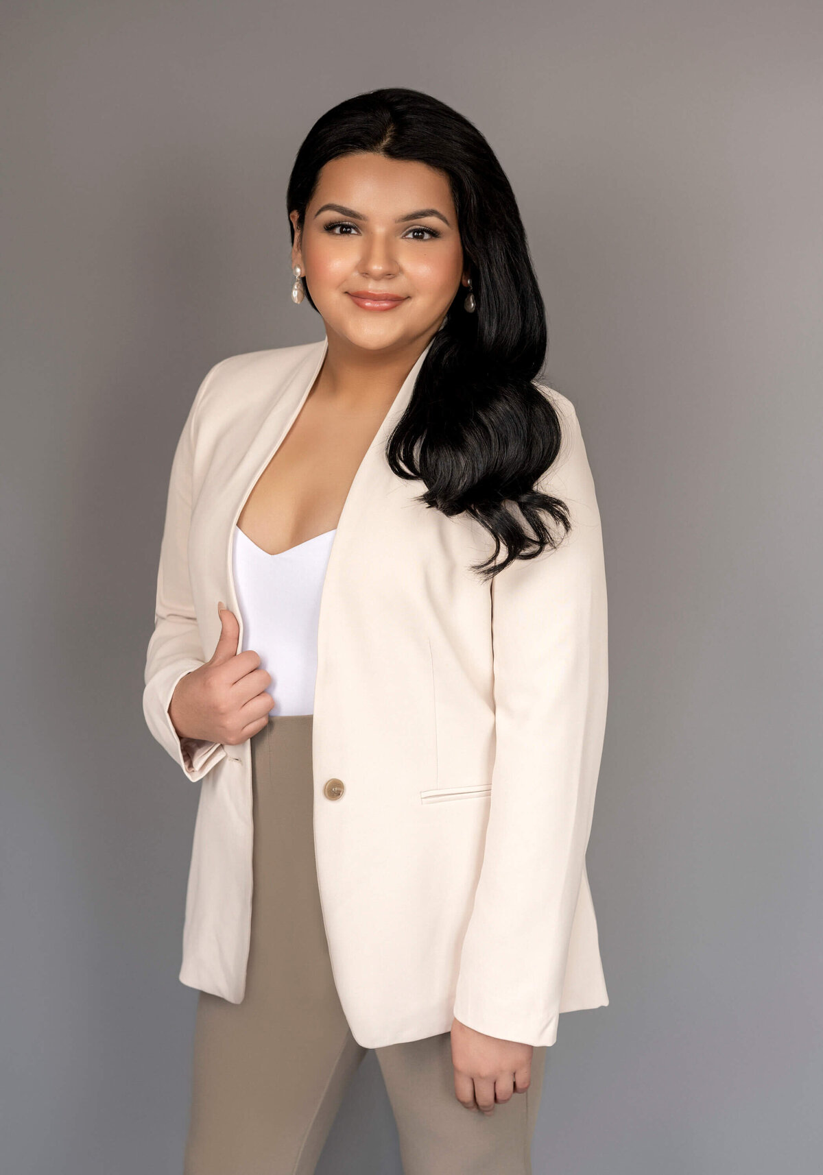 A woman in a cream blazer and fitted pants, leaning on a velvet couch, exuding professionalism.