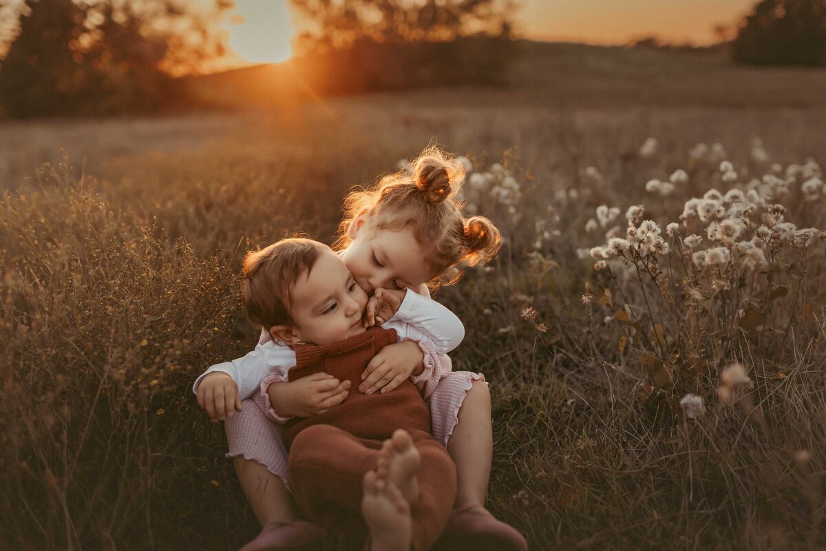 young girl holds her baby brother between her legs while looking at him sweetly. In the background is the orange Texas sunset and around them white wildflowers and grass