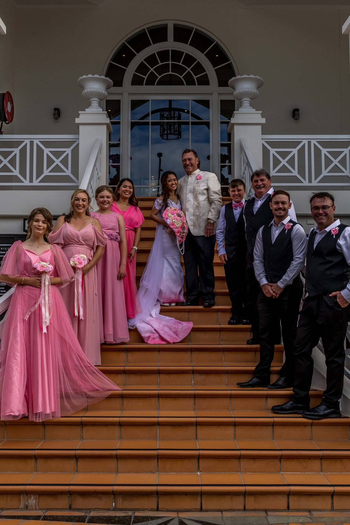 A joyful wedding party poses together on the steps of a grand building, capturing a moment of celebration and love - photo by Jamie Simmons