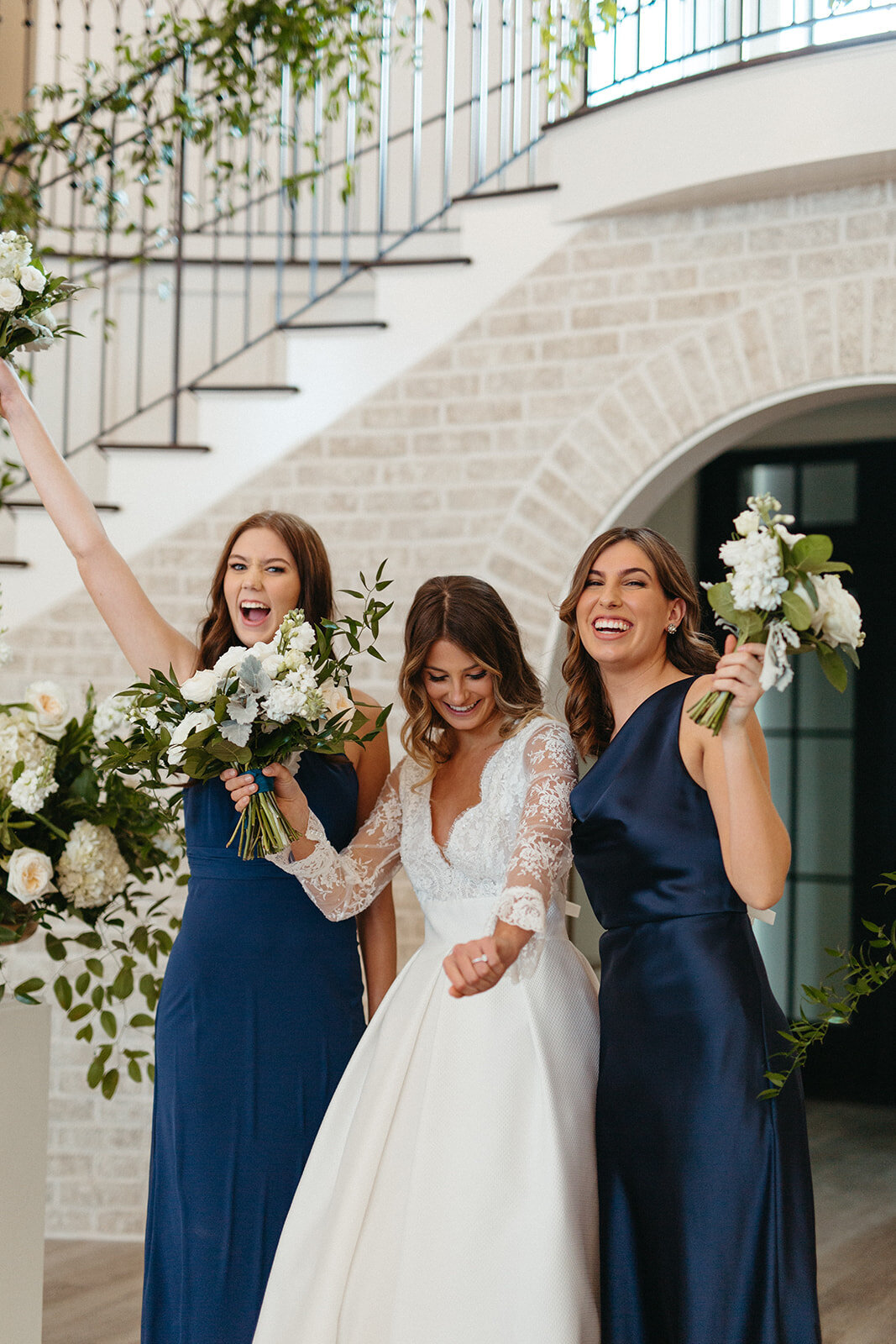 A bride in a white wedding gown and two bridesmaids in blue hold white bouquets smiling by staircase wrapped in garland.