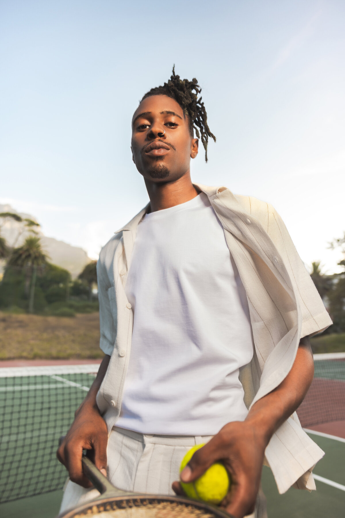 Confident man holding a tennis racket and ball on court, dressed in light tones, with Swim Essentials showcased during a golden hour session near Table Mountain.