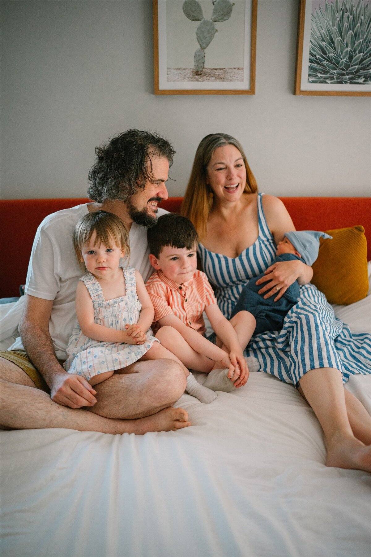 Parents sitting on a bed while one holds a newborn and the other has two small kids in their lap. 