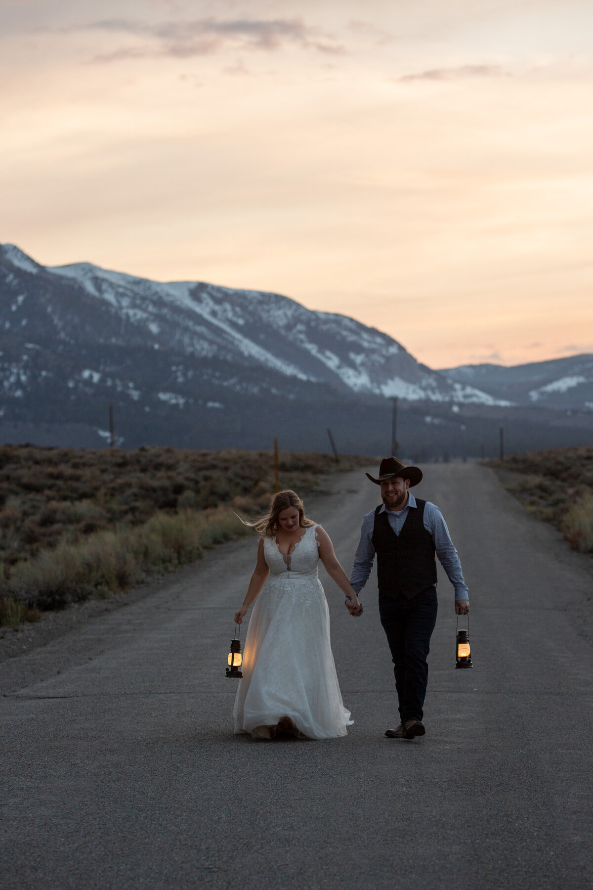 A bride and groom walk down a road in Mammoth, holding hands and holding lanterns as the sunsets behind them.