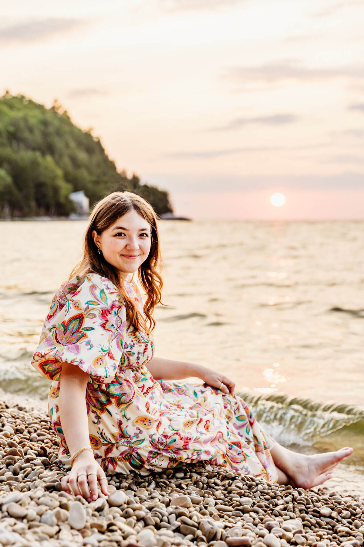 beautiful brown haired teen girl in a fun patterned dress sitting on a white stone beach as the sun sets behind her for her senior photos.