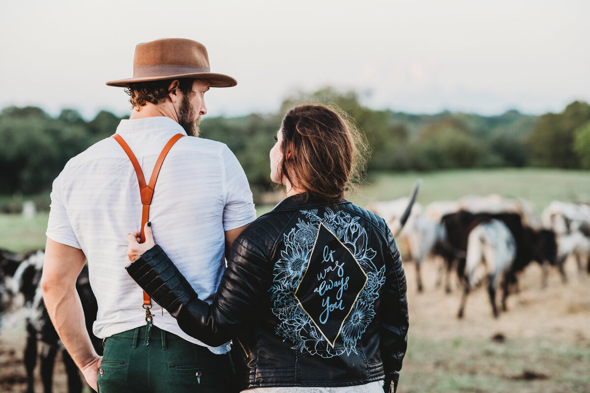 Couples Photography, Man in suspenders and a brown hat, facing longhorns, is standing next to a woman in a black leather jacket who has her hand on his back.