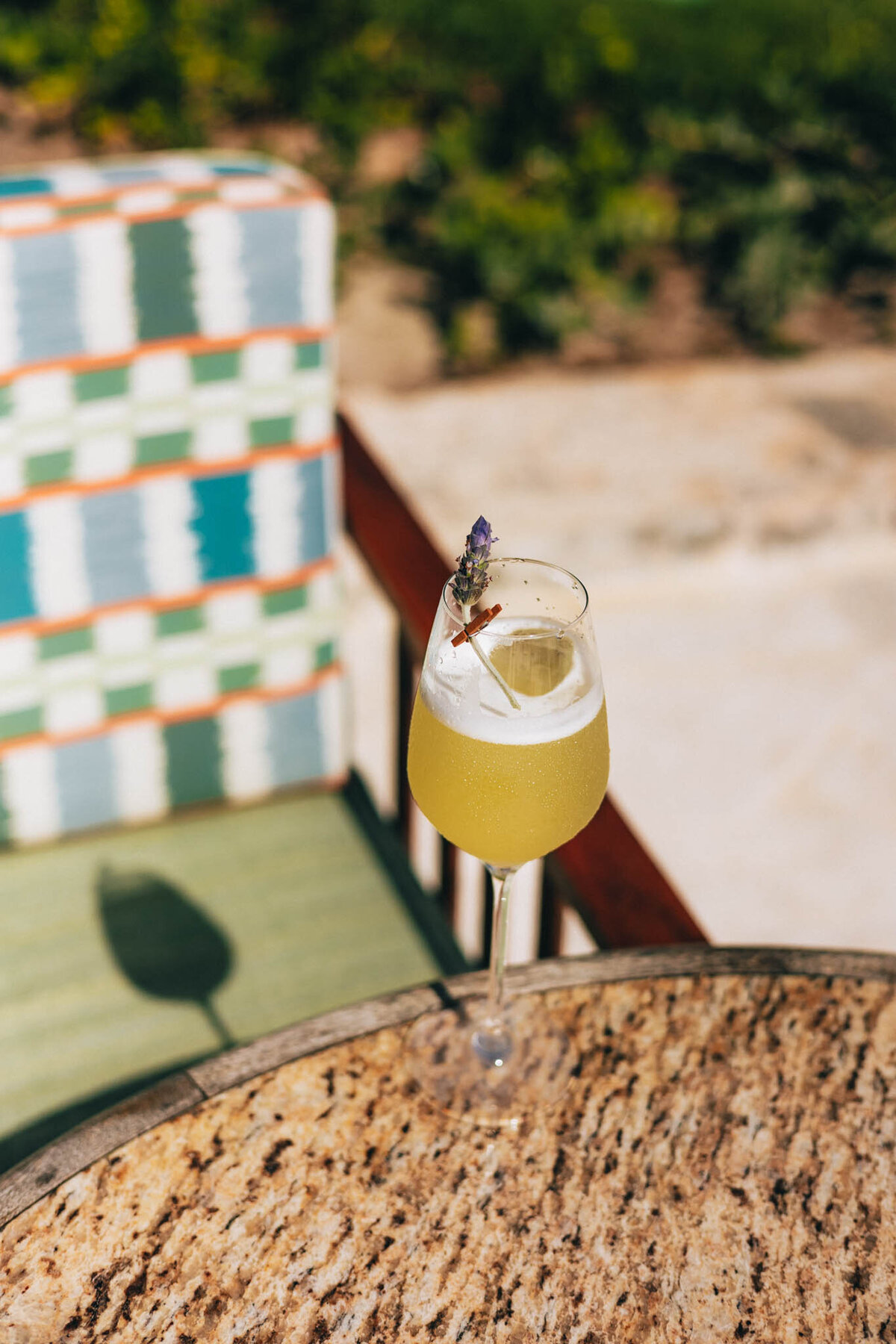 A tall glass of yellow-green cocktail garnished with a sprig sits on a round wooden table. In the background, there's a patterned chair and greenery in soft focus under bright sunlight.