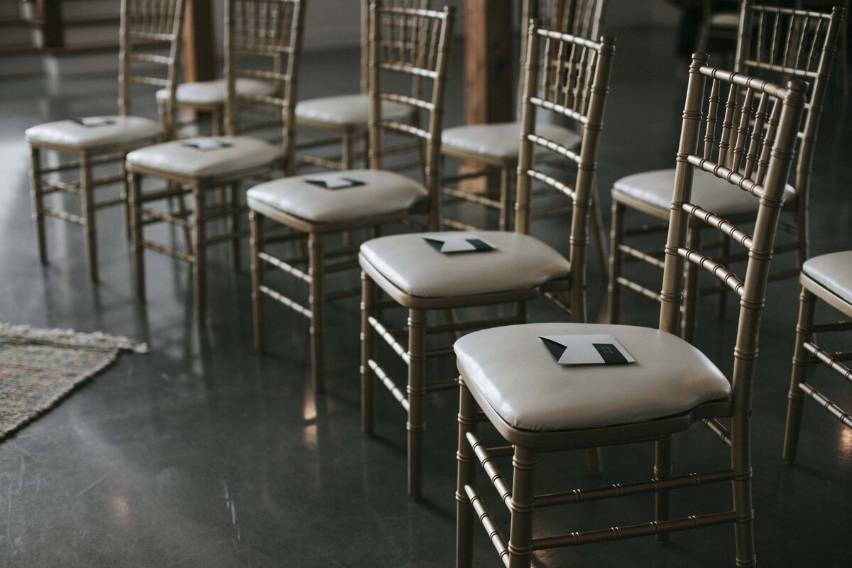 Squared white and dark green place cards are set atop gold chairs with ivory seats.