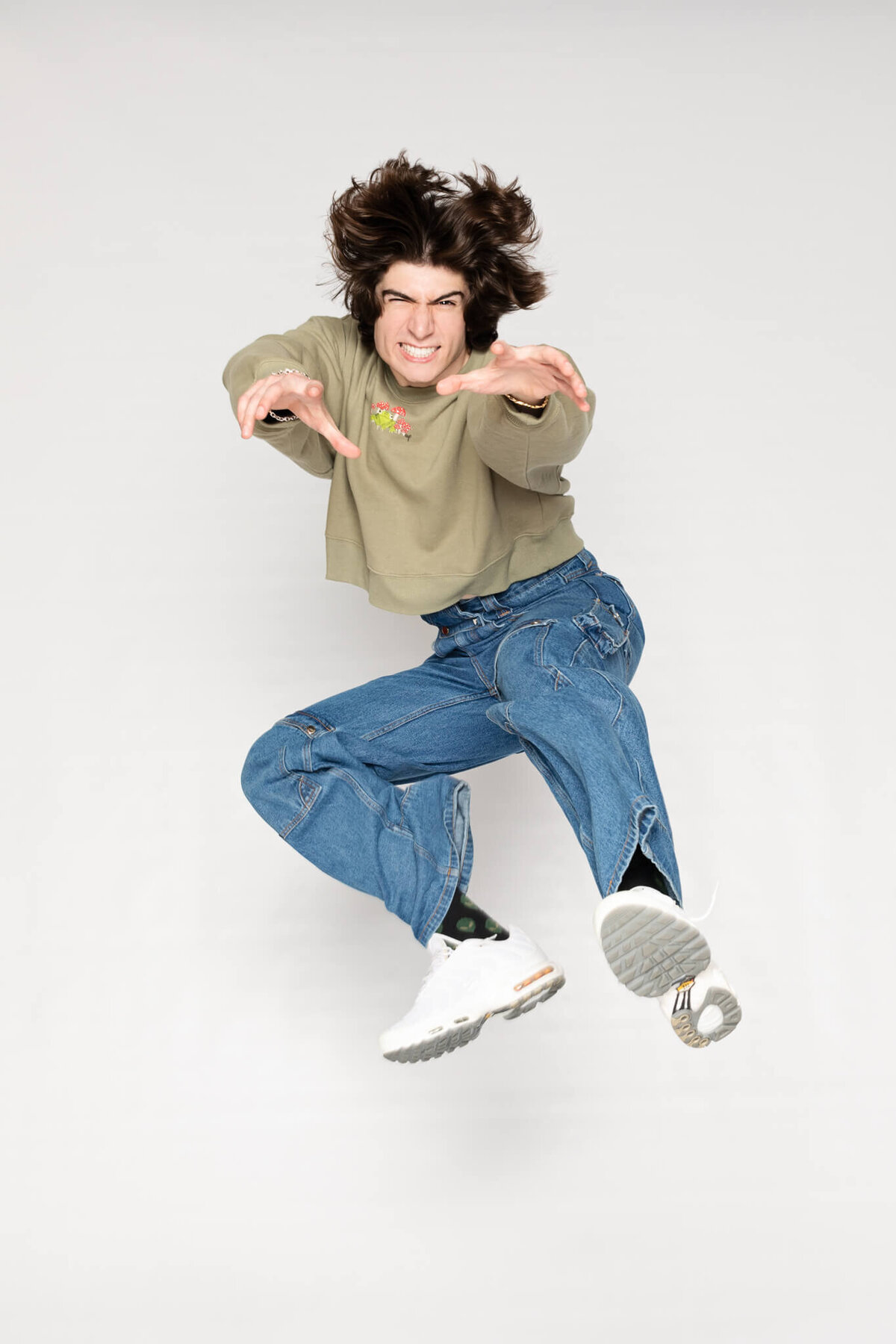 A person with long hair is mid-air and appears to be energetically jumping toward the camera. They wear a green shirt, blue jeans, and white sneakers against a plain gray background.