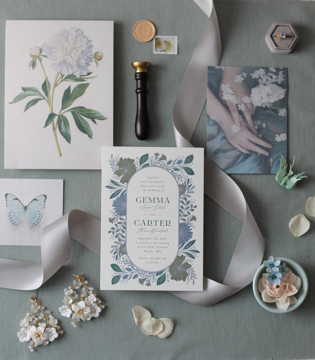 A flat lay of white blue and gray invitations, artwork, ribbon, flowers, stamps, seals, wedding jewelry, and an engagement ring with a blue stone
