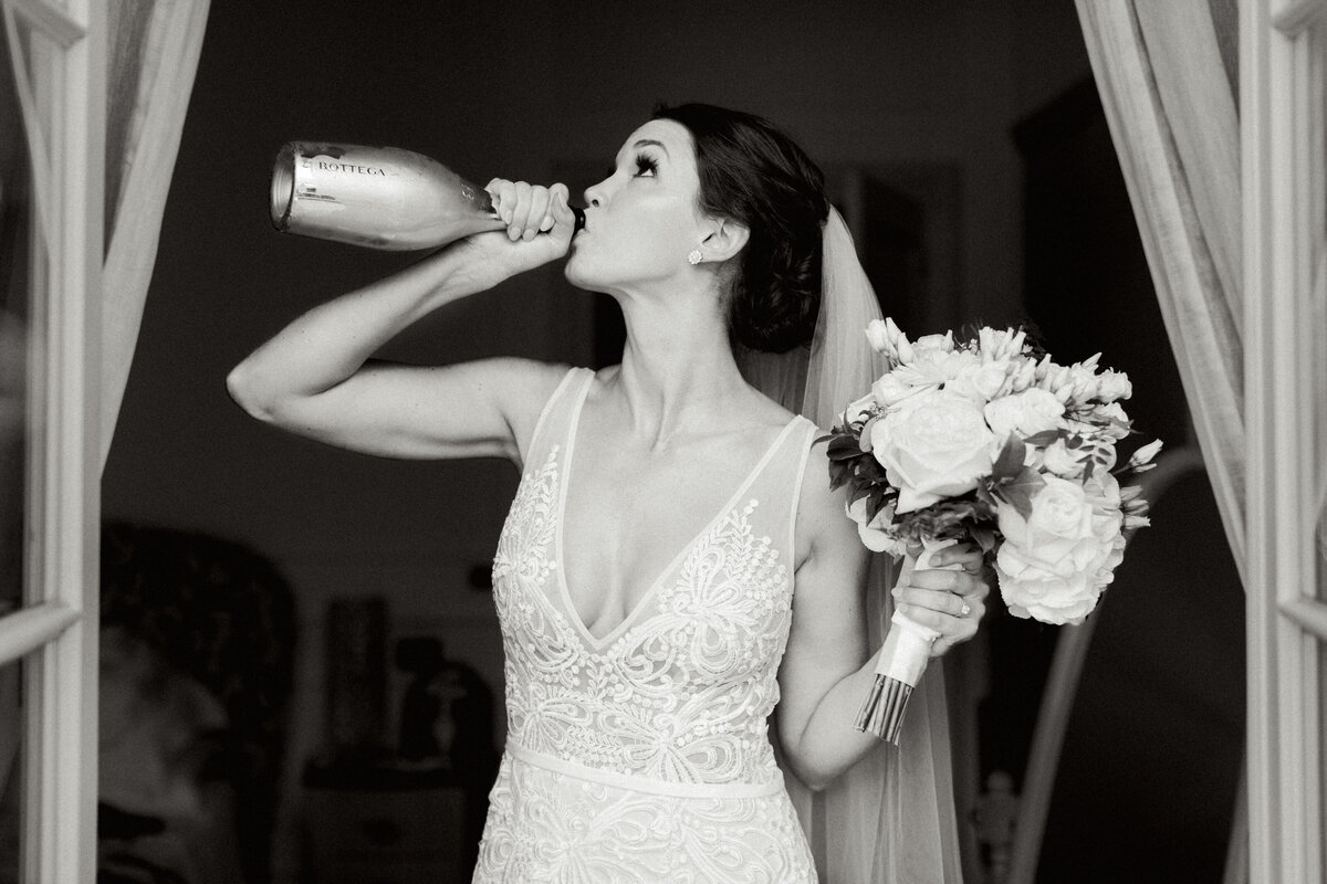 Bride holding floral bouquet and drinking bottle of wine in white lace wedding dress against black background