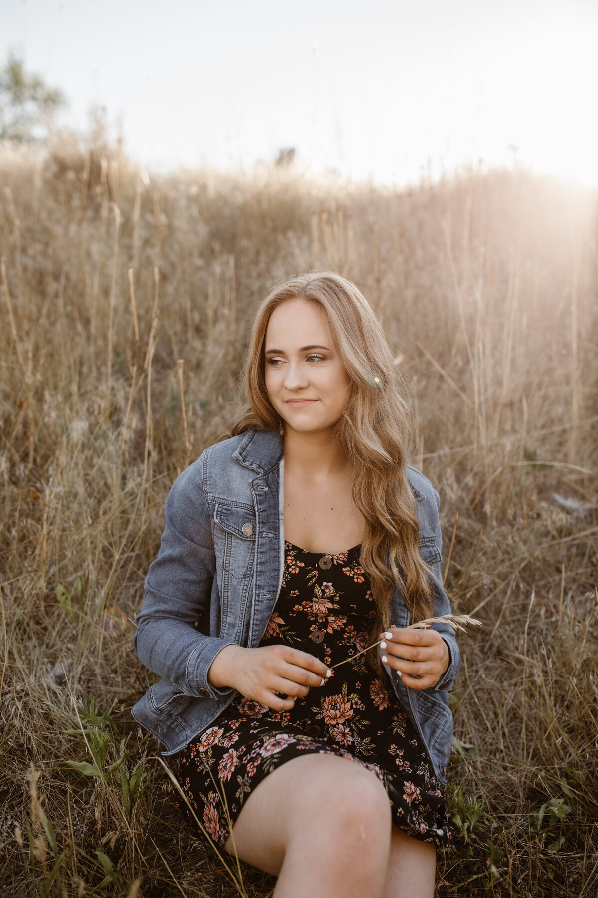 Teen girl in a floral dress and denim jacket sits in a golden wheat field at sunset, gently holding a blade of grass and smiling softly during her senior photo session.