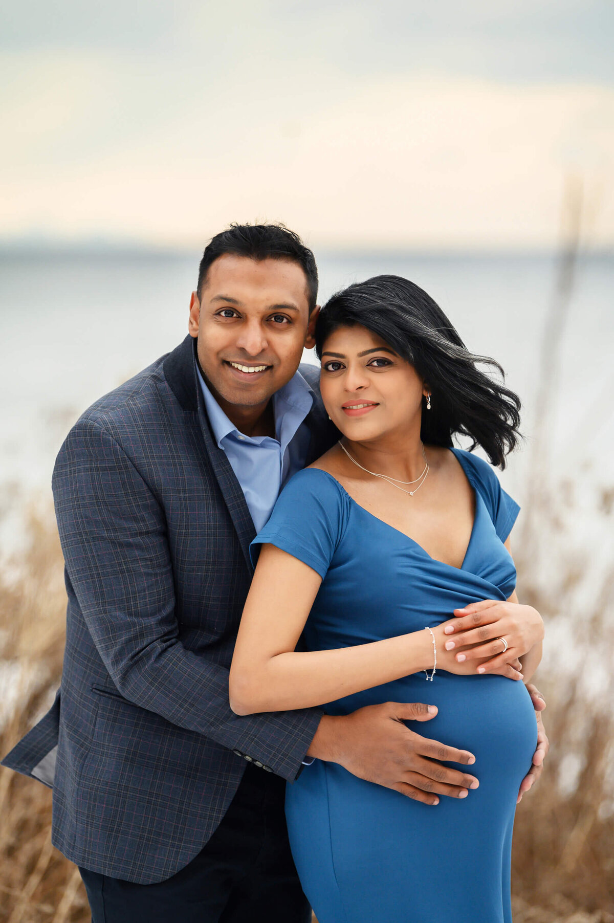 Pregnant woman wearing a blue maternity dress with her husband hugging her outdoor for her maternity photography session Toronto.
