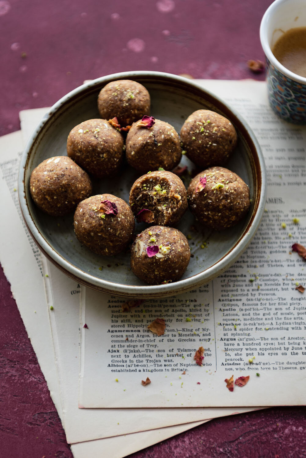 Omayah Atassi Dubai Food Photographer - 45 degree angle with energy balls in a bowl, on top of a purple backdrop and old book pages, with rose petals scattered about and a coffee cup in a corner