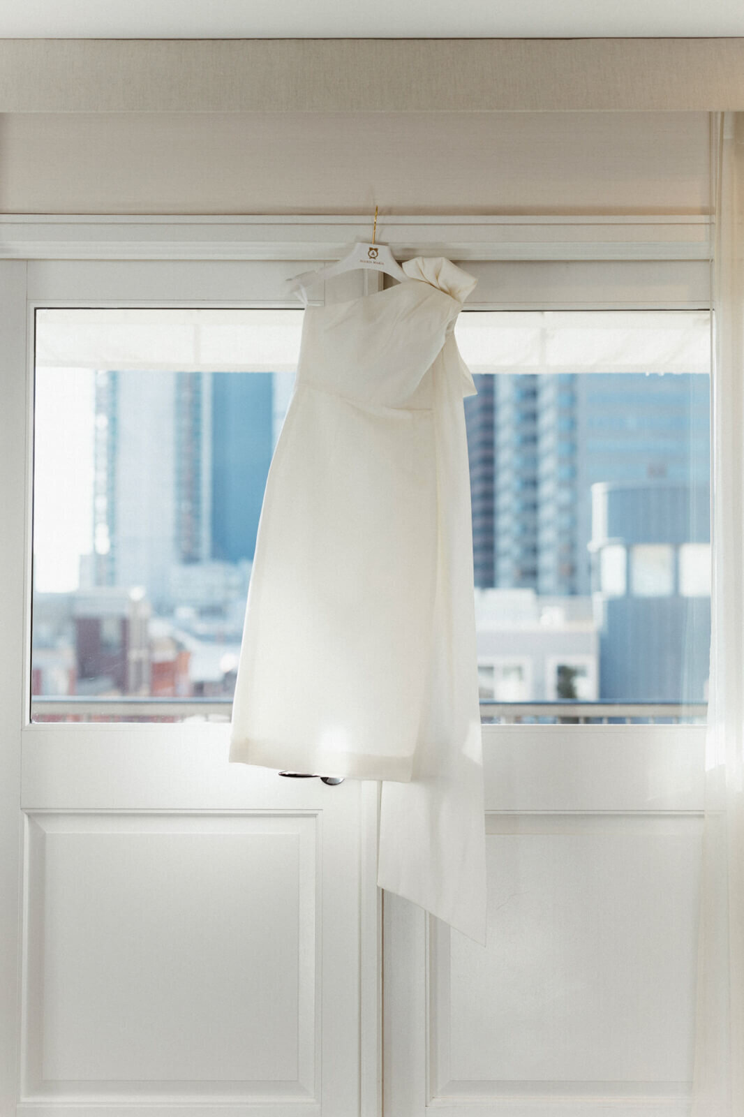 A wedding dress hanging in a window with a view of many buildings outside