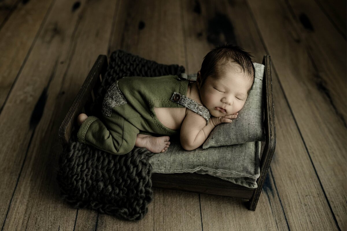 little newborn boy wearing green is posed on a little newborn baby bed in his studio newborn session